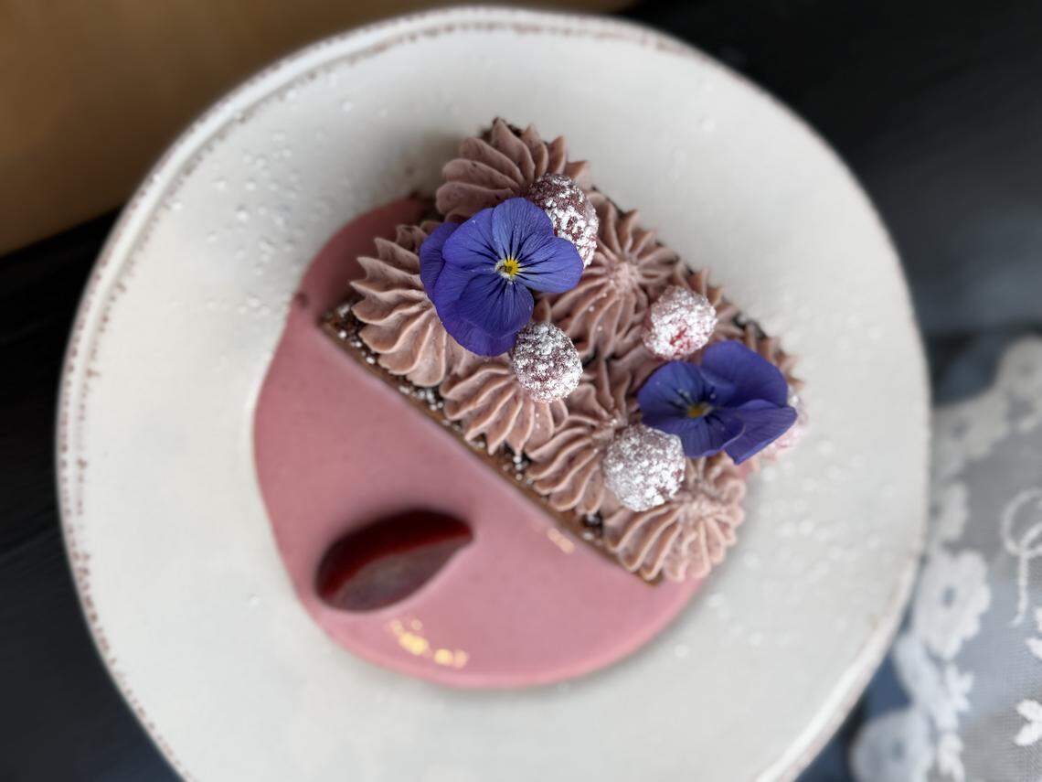 A rectangular layered dessert topped with piped pink cream, sugared raspberries, and two purple edible pansies, served on a white plate with a dark pink sauce.