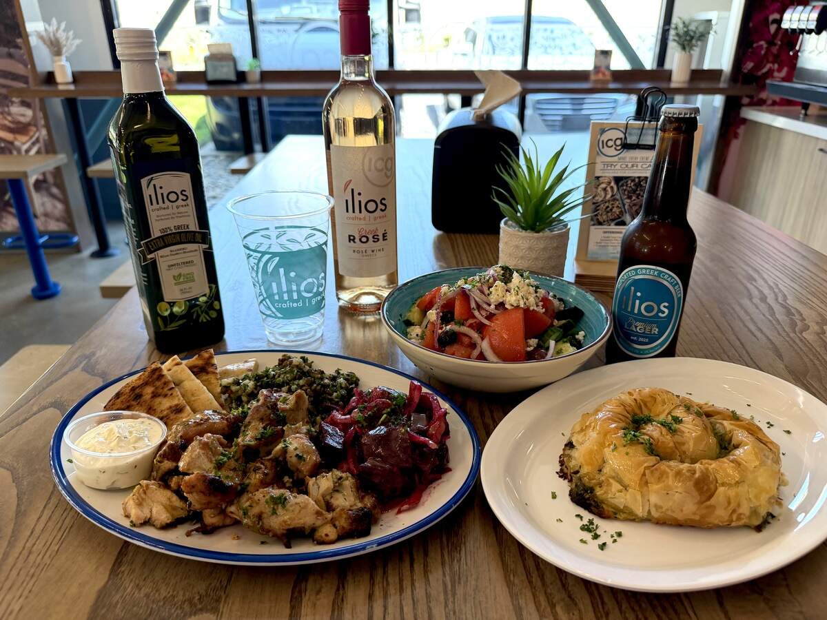 A spread of Greek dishes on a wooden table, featuring a platter of grilled chicken, pita, and dipping sauce, a bowl of tomato salad, and a plate with a golden pastry roll, flanked by bottles of Ilios wine, olive oil, and craft beer.
