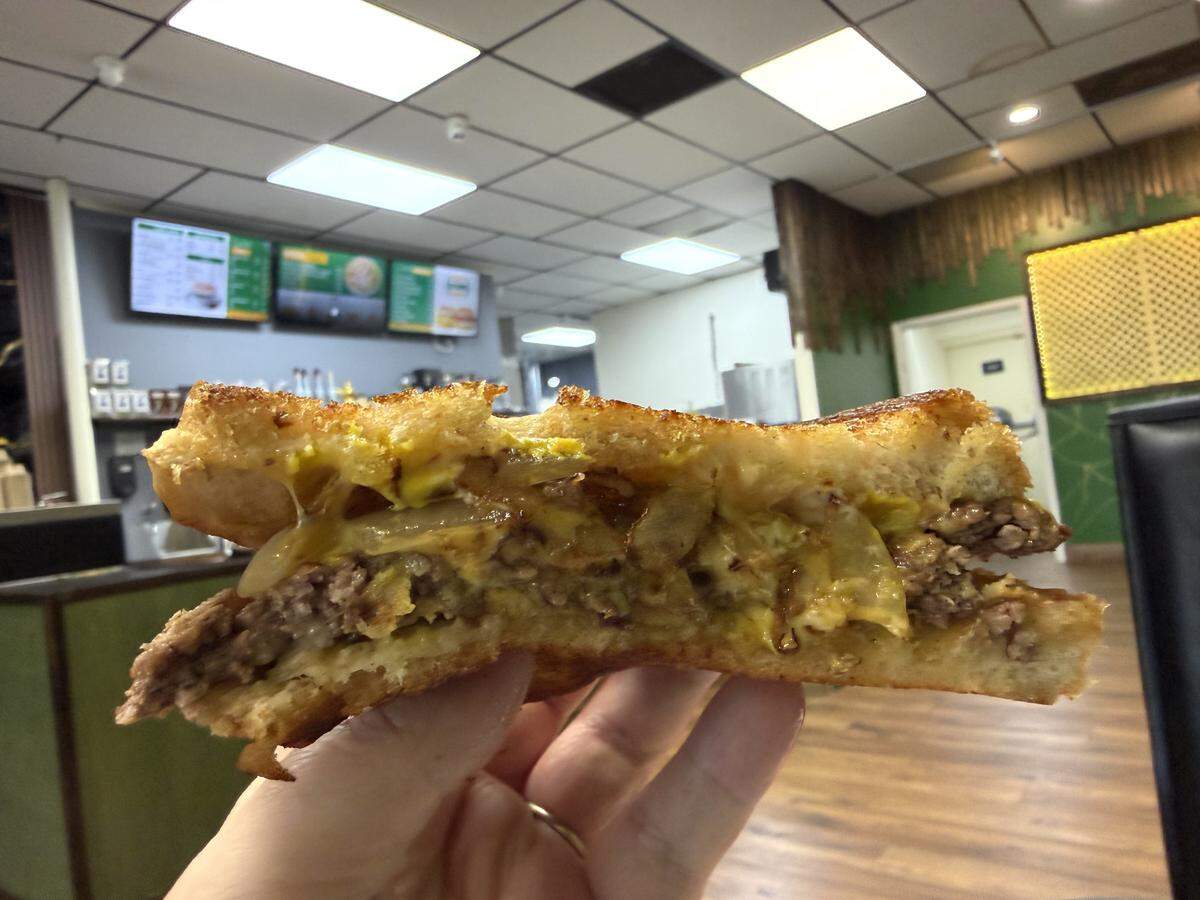A close-up shot of a hand holding a cross-section of a freshly cut patty melt inside a restaurant. The sandwich features a seasoned beef patty, melted cheese, and grilled onions nestled between two slices of golden-brown, toasted bread. The background is a softly blurred restaurant interior showing booth seating, a service counter, and digital menu boards hanging on the wall.