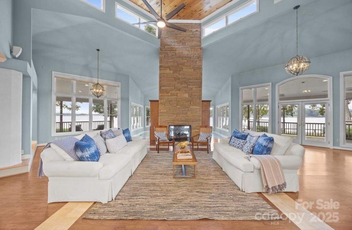 A wide-angle interior view of a spacious living room with high, vaulted ceilings painted light blue, a tall, floor-to-ceiling stone fireplace in the center, and wood floors. Two matching white sofas face each other, separated by a coffee table and sitting on a textured area rug. The room has multiple large windows and doors offering a panoramic view of a lake and outdoor scenery.