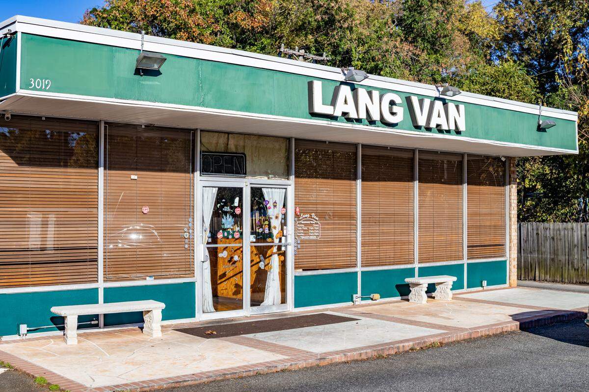 A straight-on, eye-level, wide shot of the “Lang Van” restaurant exterior on a bright, sunny day. The one-story building features a prominent green fascia with “LANG VAN” in large, white letters. The facade consists of floor-to-ceiling windows, which are covered from the inside by brown wooden blinds. A small neon “OPEN” sign is visible in the central glass-door entrance. Two simple, white stone benches sit on the concrete patio out front, and tall trees are visible in the background.