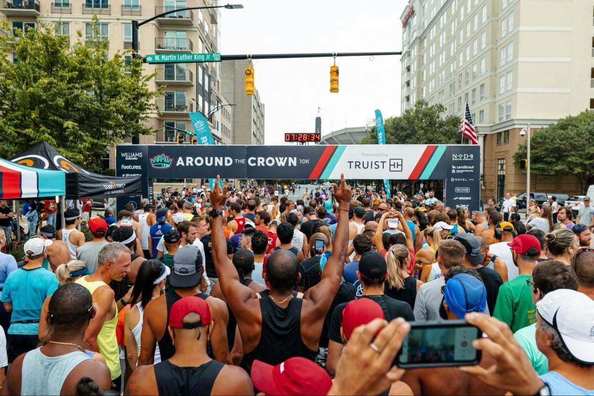 A large crowd of runners seen from behind, facing the starting line banner for the “Around the Crown 10K” race on a city street.