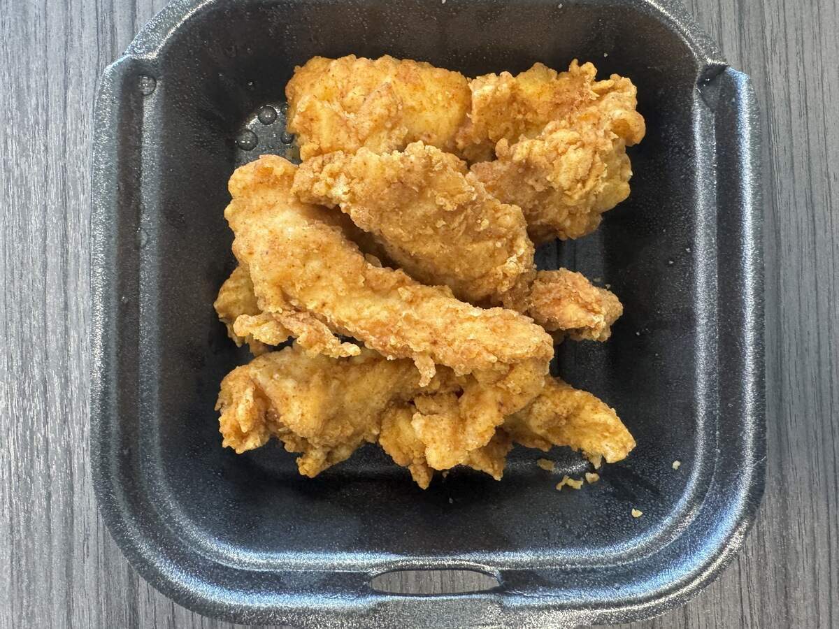 A top-down view of several fried chicken tenders neatly arranged inside an open, small black styrofoam container. The chicken strips have a deeply golden, textured, and crunchy batter and fill the bottom of the square container.