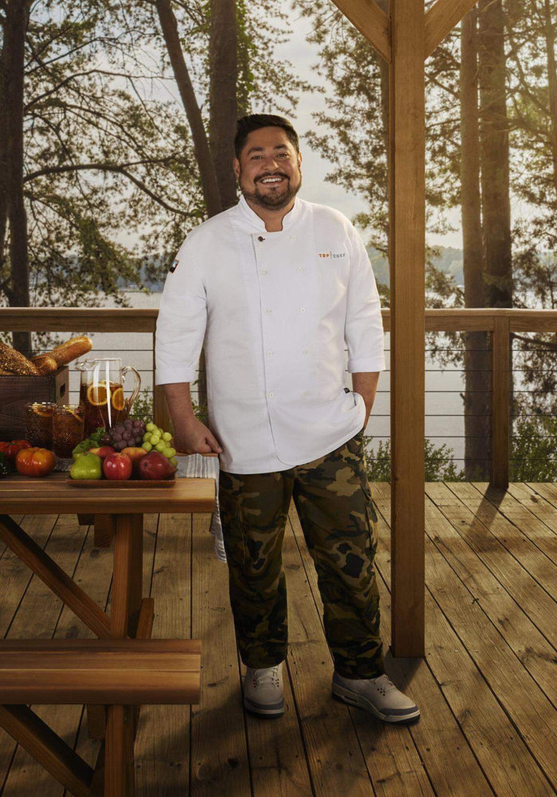 A portrait of a smiling chef wearing a white chef’s coat with “Top Chef” embroidered on the chest and standing outdoors on a wooden deck with a scenic background of trees and water. In the foreground, a display of fresh food includes loaves of bread, a pitcher of iced tea with orange slices, apples, and grapes.