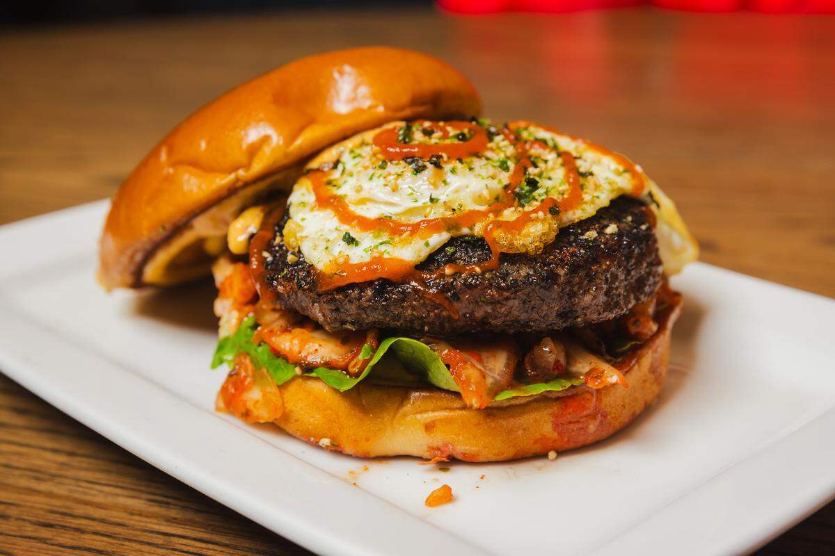 A close-up of a specialty burger served on a white rectangular plate. The burger features a thick, charred beef patty topped with a fried egg, drizzled with red sriracha-style sauce and green garnishes. A generous layer of red kimchi and fresh lettuce sits beneath the patty on a toasted brioche bun.