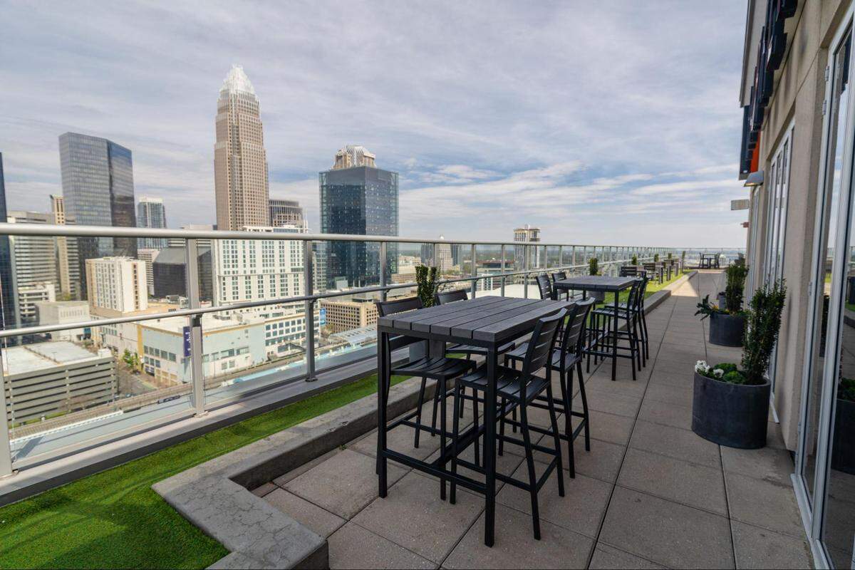 A panoramic shot of an urban rooftop patio with a stunning view of a city skyline. In the foreground, several black high-top tables with matching chairs are arranged on a paved and artificial grass surface. A glass and metal railing runs along the edge of the patio, offering an unobstructed view of the city. The skyline features multiple tall buildings and skyscrapers under a cloudy blue sky.
