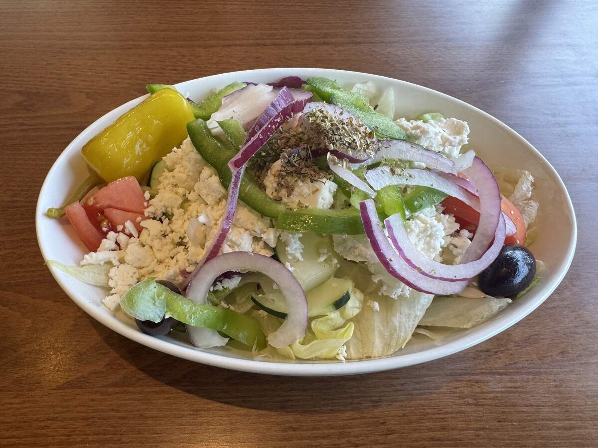 A close-up, eye-level shot of a Greek salad served in a white oval bowl on a wooden table. The salad consists of chopped iceberg lettuce topped with crumbled feta cheese, sliced green bell peppers, red onion rings, cucumber slices, tomato wedges, a yellow pepperoncini, black olives, and a sprinkle of dried oregano.