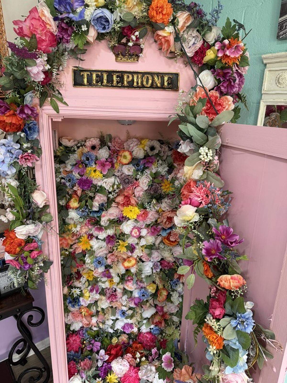 A light pink, English-style telephone booth is shown. The booth’s interior is completely filled with a colorful wall of artificial flowers in various shades of pink, purple, yellow, orange, and blue. A black and gold sign above the booth’s open door reads “TELEPHONE.” The outside of the booth is also decorated with colorful floral garlands.