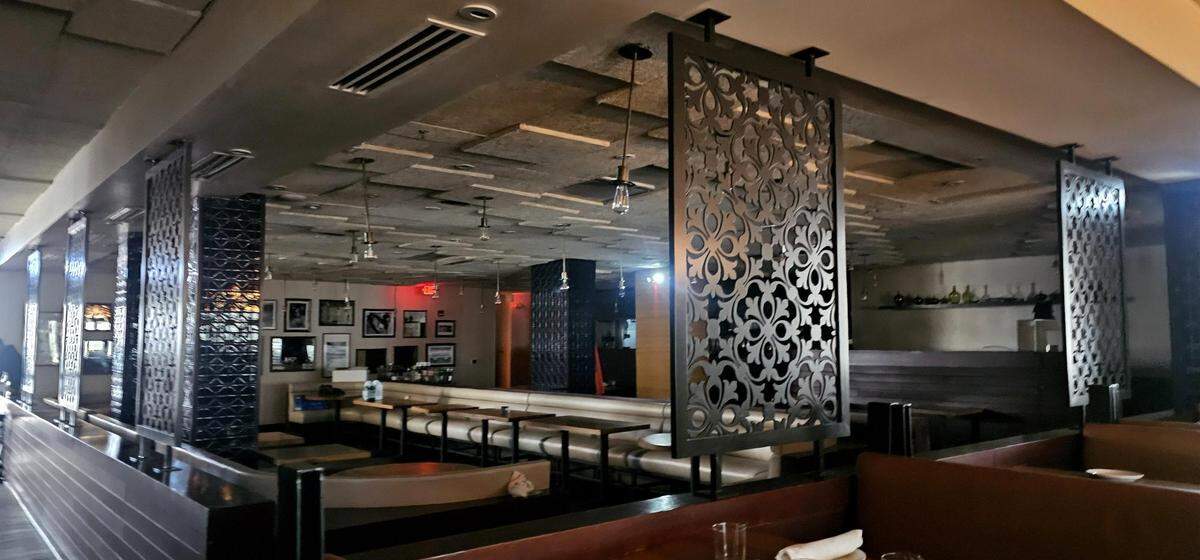 A wide, interior shot of a restaurant dining room featuring low lighting and modern decor. The space is divided by decorative, perforated metal screens and columns. Booth seating lines the perimeter, and the ceiling has textured, geometric sound dampening panels.