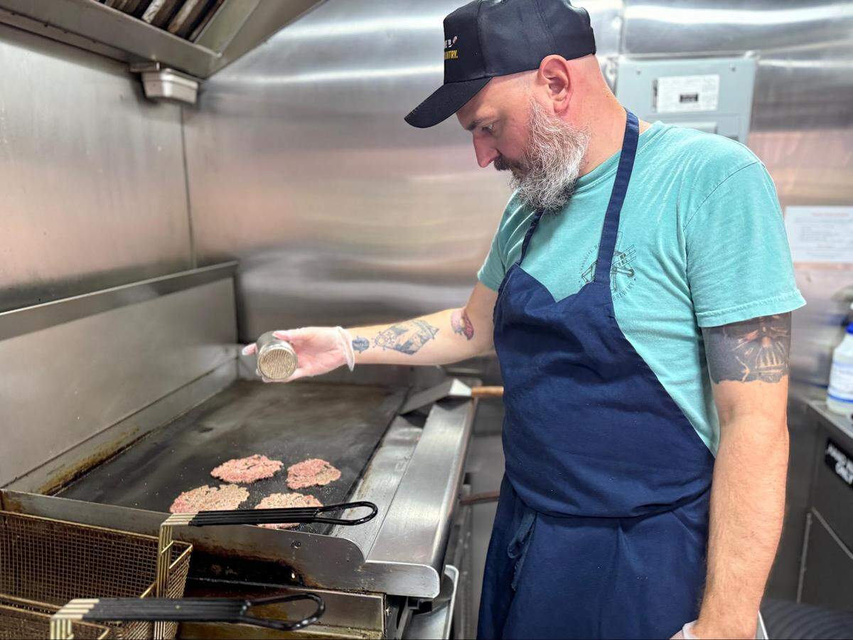 A chef with a beard, a black baseball cap, and tattoos on their arms is standing at a flattop grill, sprinkling seasoning onto several burger patties. They are wearing a blue apron and gloves. The kitchen area is made of stainless steel.