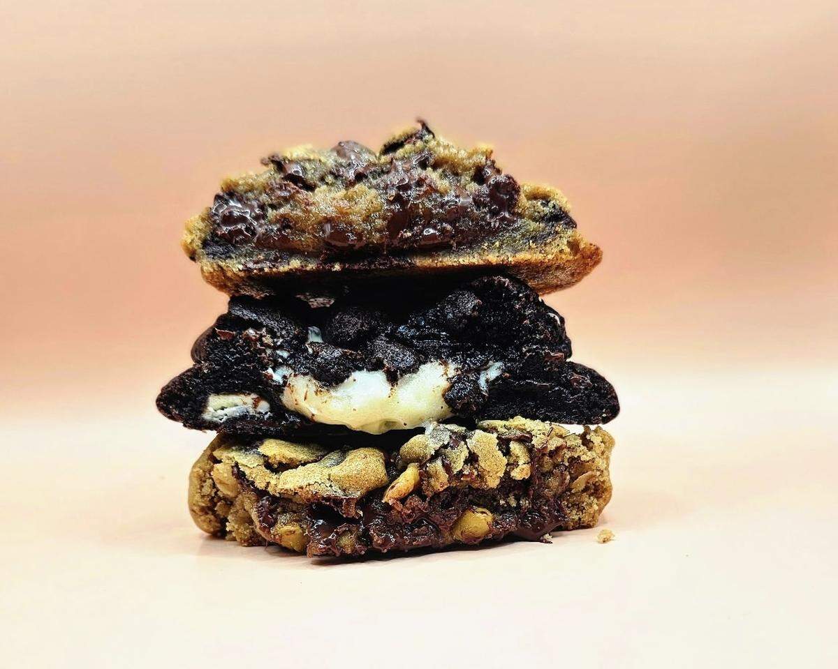 A professional food photograph of three gourmet cookies stacked vertically on a peachy-pink surface. Each cookie is broken in half to showcase its texture: a gooey chocolate chip on top, a cookies-and-cream style cookie in the middle, and a loaded oatmeal-nut cookie on the bottom.