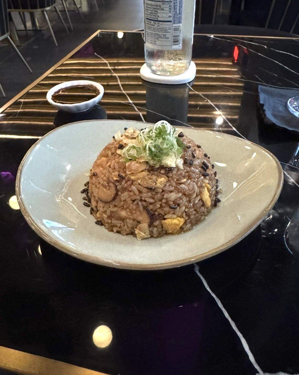 A dome-shaped serving of fried rice topped with chopped scallions sits on a light gray plate. The rice appears to include egg and pieces of lobster or seafood. A small dish of soy sauce and a bottle of sparkling water rest on a glossy black marble table in the background.