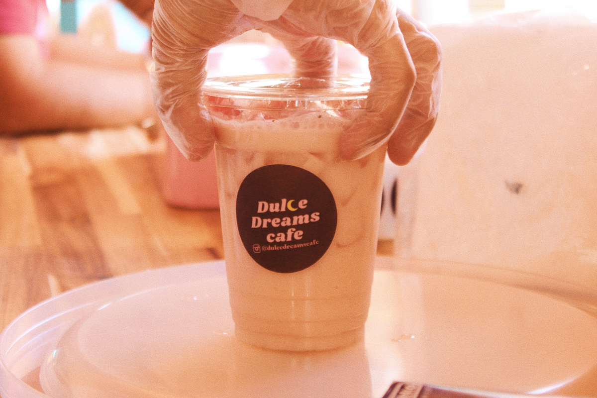 A hand wearing clear plastic gloves holds a clear plastic cup filled with a pale, creamy iced drink. A black circular sticker on the cup reads “Dulce Dreams cafe” with the handle “@dulcedreamscafe” underneath. The background is a soft, warm blur.