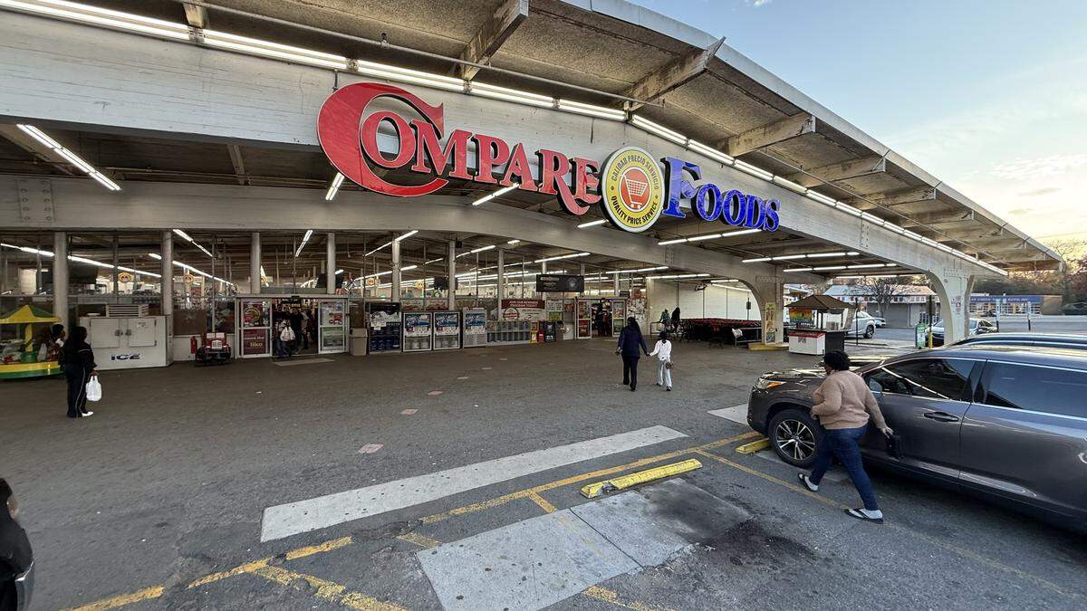 Exterior, wide shot of a Compare Foods supermarket entrance with a large red and blue sign. The parking lot in front is mostly empty, and a few people are walking towards or away from the entrance.