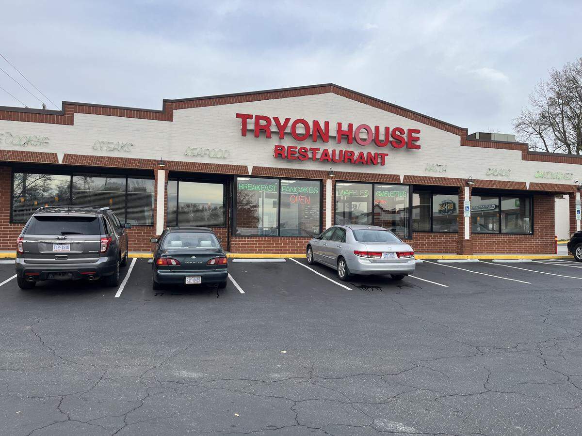 The exterior of the Tryon House Restaurant, a long, brick and beige building with a central gable displaying the name in large red letters. Three cars are parked in the asphalt lot in front of the large windows.