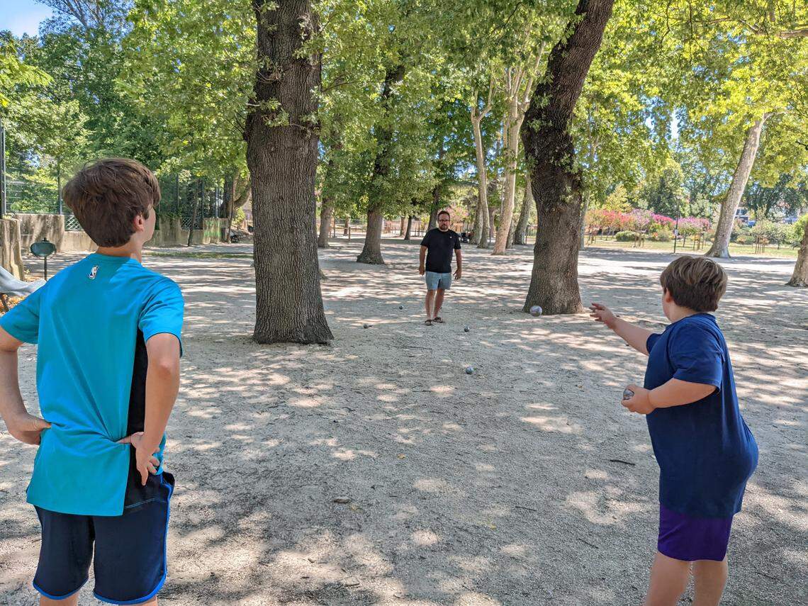 From a perspective behind two young children, a father is seen in the distance as they all play a game of pétanque. The boy on the right is throwing a silver ball, while the boy on the left has his hands on his hips. The game takes place on a gravel court with dappled sunlight filtering through the trees.
