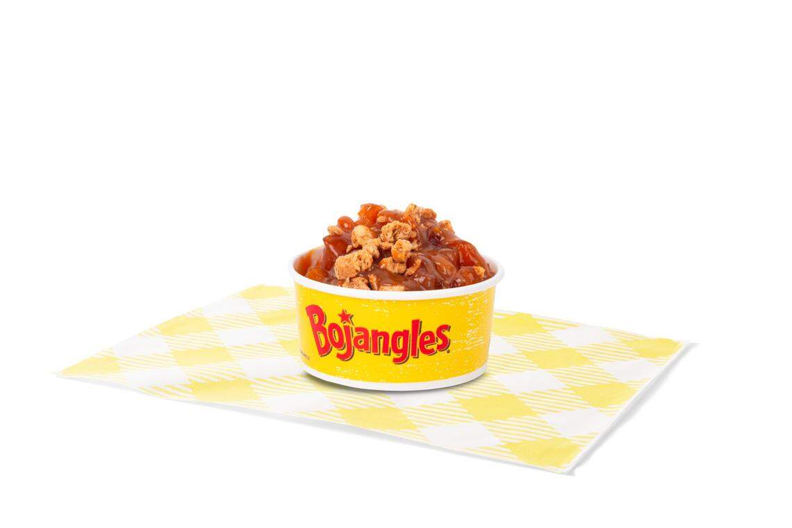 A bright yellow Bojangles cup filled with a Salted Caramel Apple Crisp sits on a yellow and white checkered paper square. The dessert is topped with a generous amount of cinnamon-spiced streusel crumble and a glossy caramel sauce, contrasting with the dark fruit underneath.