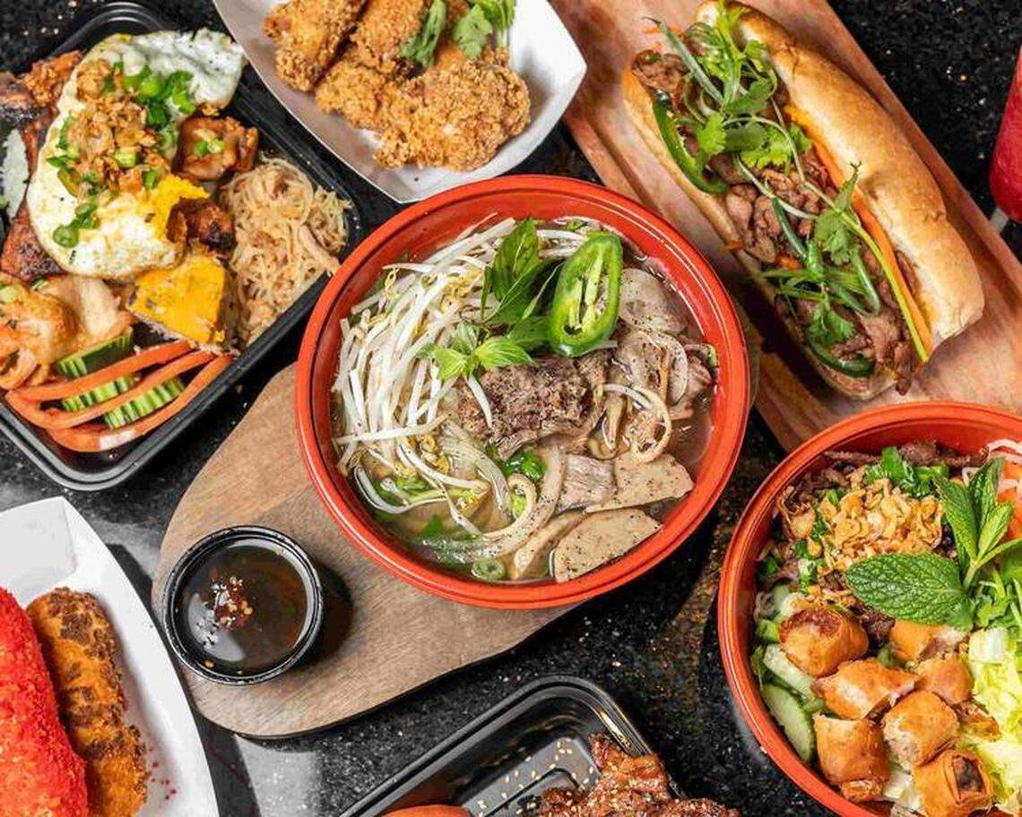 A top-down view of a diverse Vietnamese feast shows various dishes including a large bowl of beef pho with bean sprouts, a Bánh Mì sandwich, and crispy spring rolls. The spread also features fried chicken wings, a rice platter with a fried egg, and dipping sauces arranged on a dark tabletop.