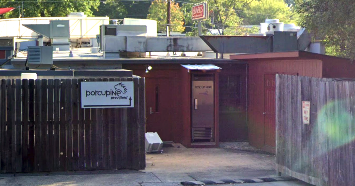 The exterior entrance of a building called “Porcupine Provision.” A white sign with the name and a stylized porcupine image is mounted on a wooden fence to the left. The entrance area is recessed, featuring a metal “PICK UP HERE” dispenser cabinet and a narrow awning. Various HVAC units are visible on the roof.