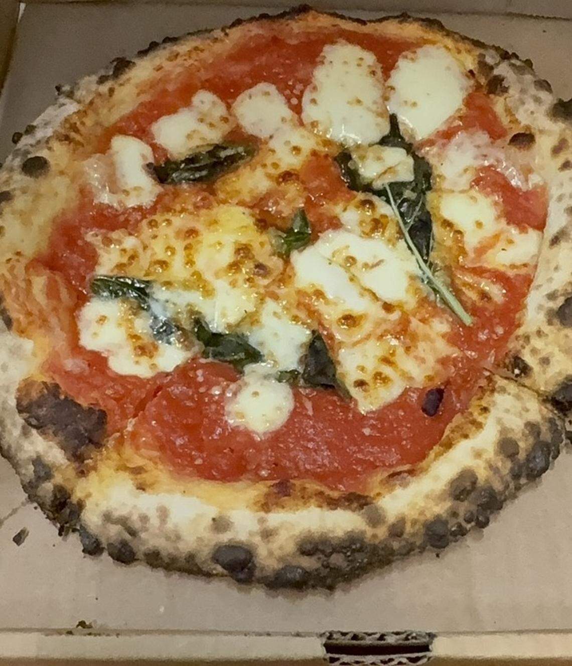 A close-up, top-down view of a personal-sized Margherita pizza inside a brown cardboard box. The pizza features a thick, charred, and bubbly “leopard-spotted” crust typical of wood-fired ovens. It is topped with a vibrant red tomato sauce, large melted dollops of fresh white mozzarella cheese, and several wilted green basil leaves.