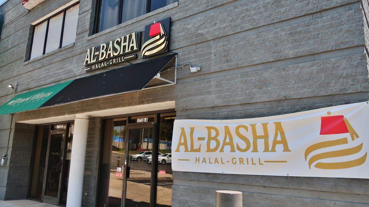 An angled, eye-level shot of the exterior of Al-Basha Halal Grill, located in a gray stone building. The restaurant features a black awning and a sign with gold lettering and a red fez icon. A large white vinyl banner with the same branding is stretched across the wall to the right of the glass entrance doors. To the left, a green awning for an adjacent business is partially visible.