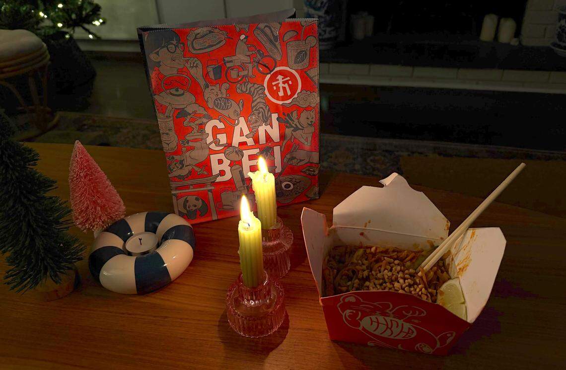 A dimly lit still-life setting on a wooden table, featuring a takeout box of noodles with chopsticks, a large red “GAN BEI” paper bag from Hawkers, two lit candles in glass holders, a small green pine tree decoration, and a blue and white striped ring-shaped object.