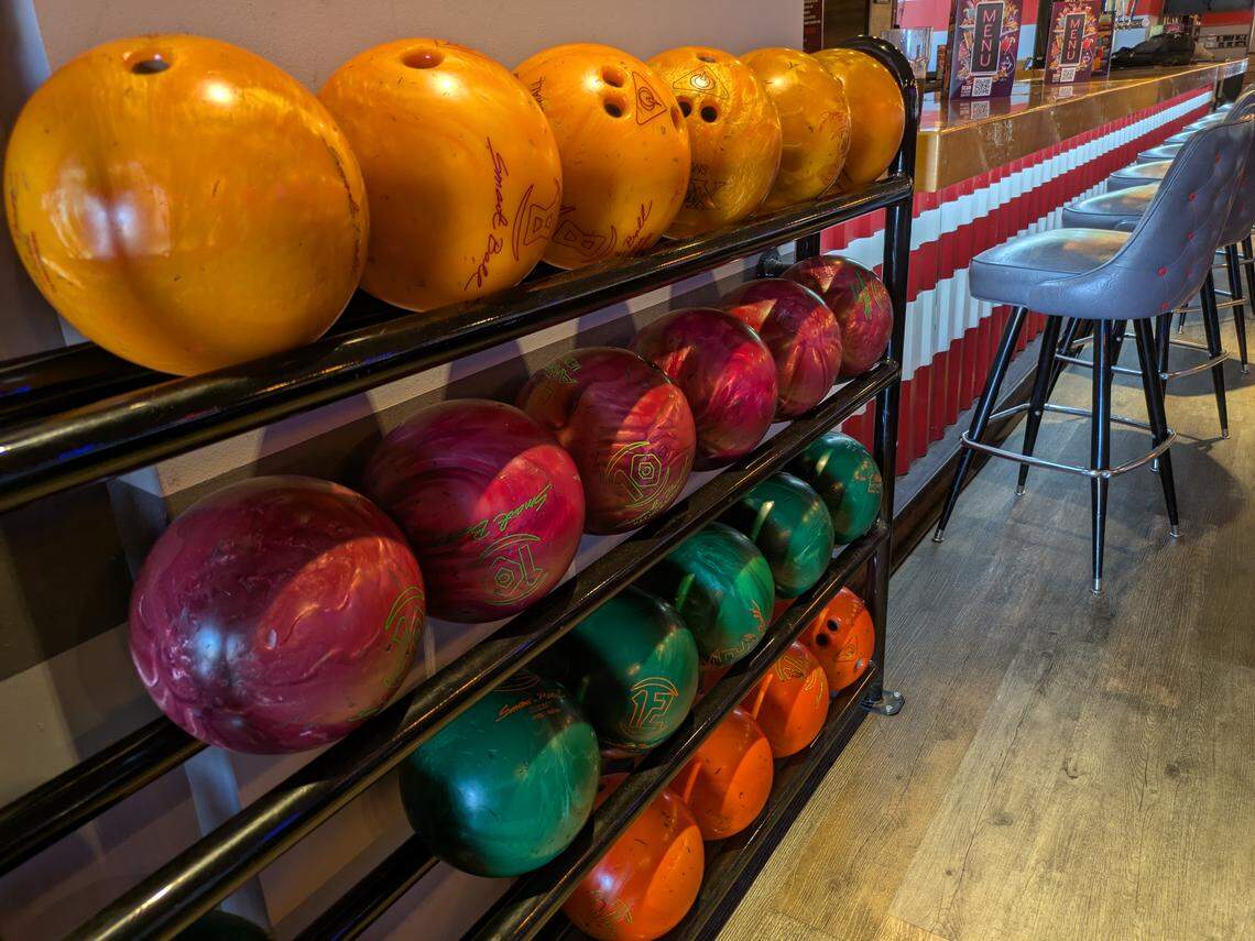 A close-up, angled shot of a four-tiered black metal rack holding numerous colorful bowling balls. The balls are organized by color, with a top row of yellow and orange, a second of red and purple, a third of green, and a bottom row of orange. To the right, the bar area is visible, featuring grey barstools lined up at a counter with a red and white striped base.