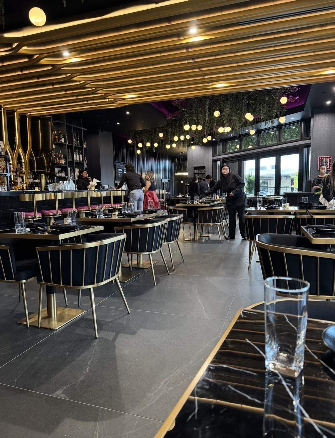 The elegant interior of a modern restaurant, featuring black and gold tables and chairs, a dark bar area, and a ceiling with glowing linear light fixtures.