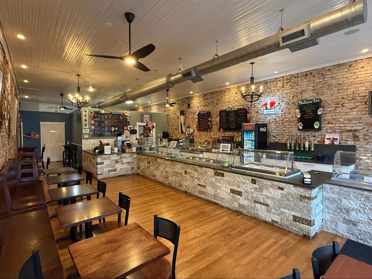 A wide interior shot of Waxhaw Creamery featuring an industrial-chic design with exposed brick walls, a long stone-tiled service counter, and wooden floors. Small wooden tables and chairs line the left side under warm chandelier lighting.