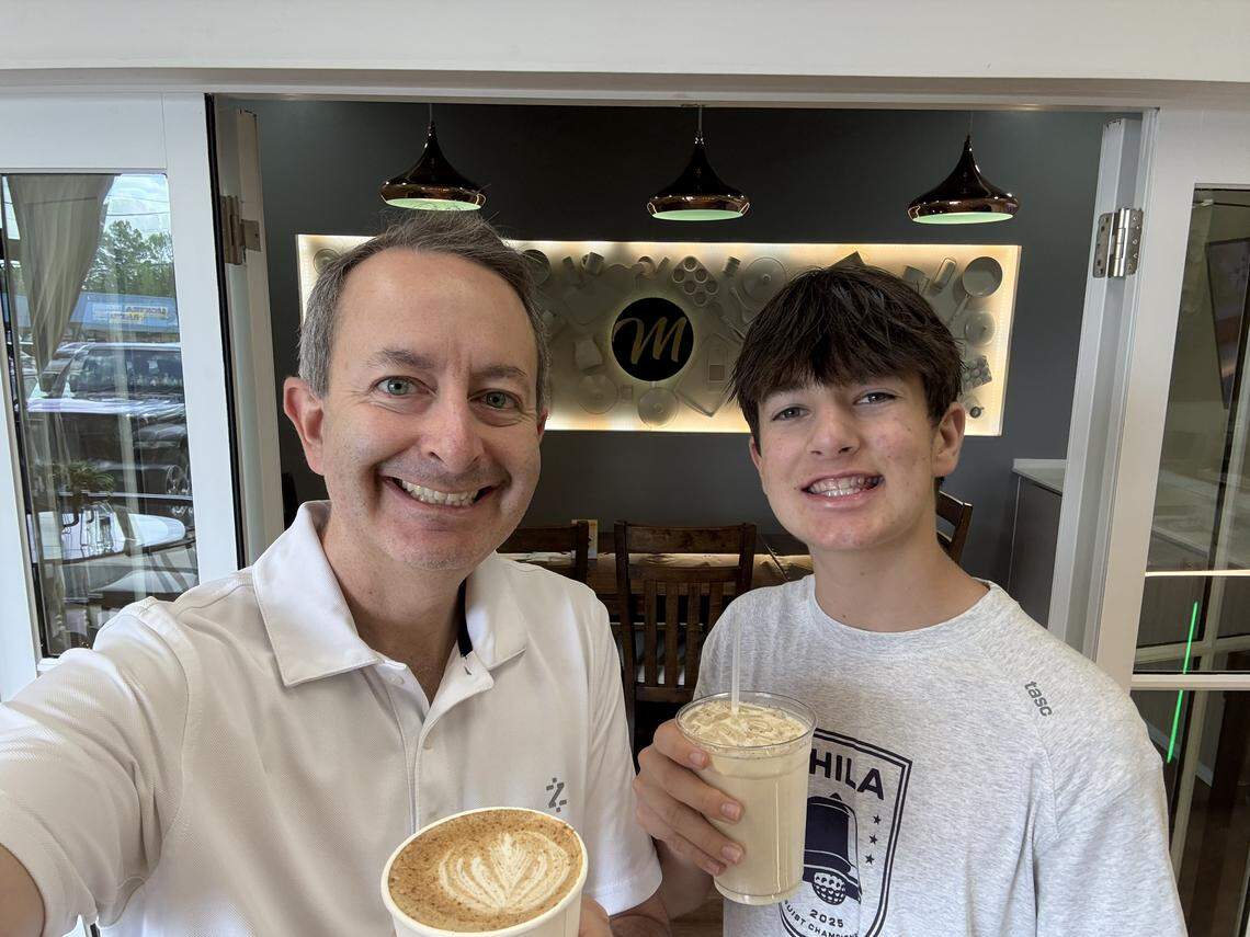 A selfie of an adult and teenager smiling while holding their coffee drinks. They are standing in the cafe with the “M” logo and backlit kitchen utensil wall display clearly visible in the background between them.
