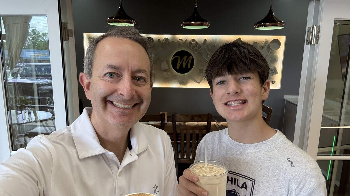 A selfie of an adult and teenager smiling while holding their coffee drinks. They are standing in the cafe with the “M” logo and backlit kitchen utensil wall display clearly visible in the background between them.