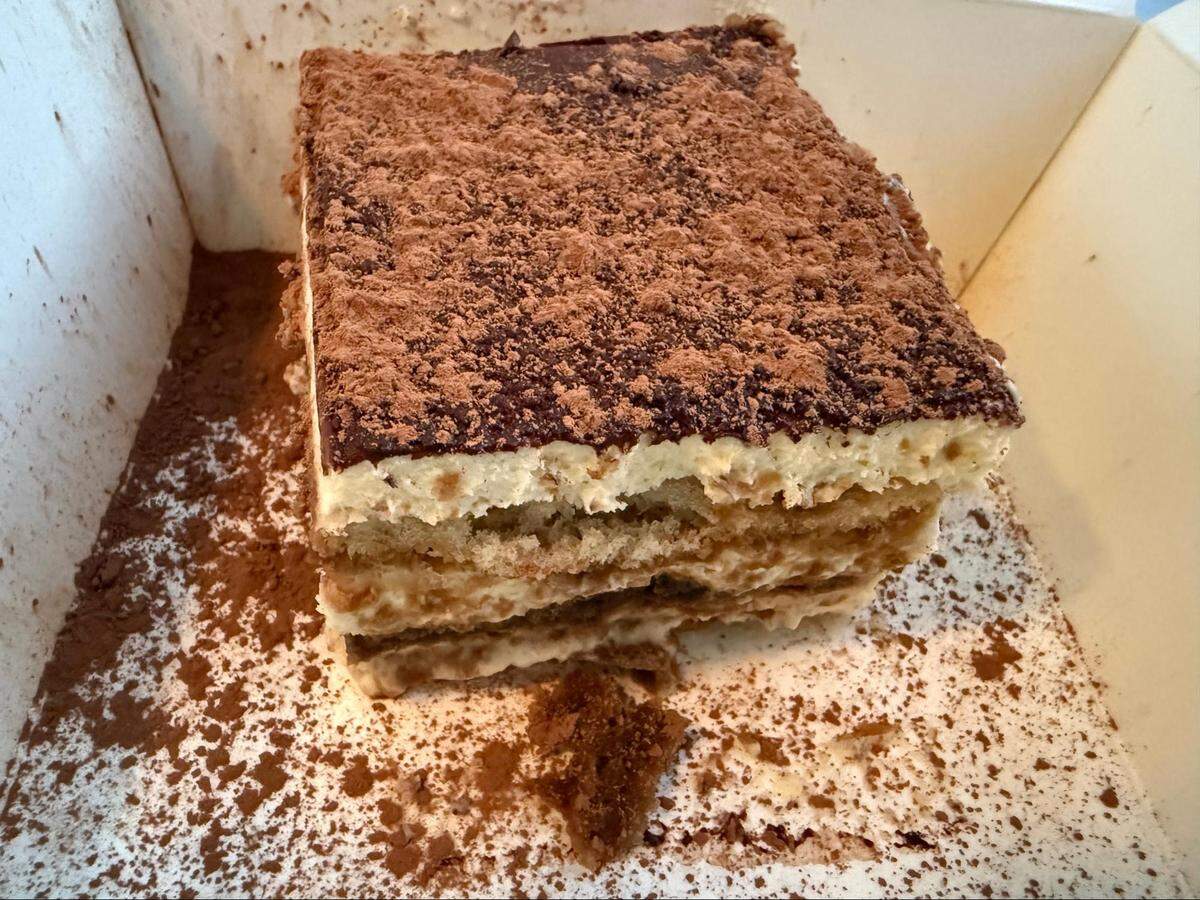 A high-angle, close-up shot of a square slice of tiramisu sitting in a white takeaway box. The dessert shows layers of cream and coffee-soaked cake, and its top is generously dusted with cocoa powder, which has also spilled onto the bottom of the box.