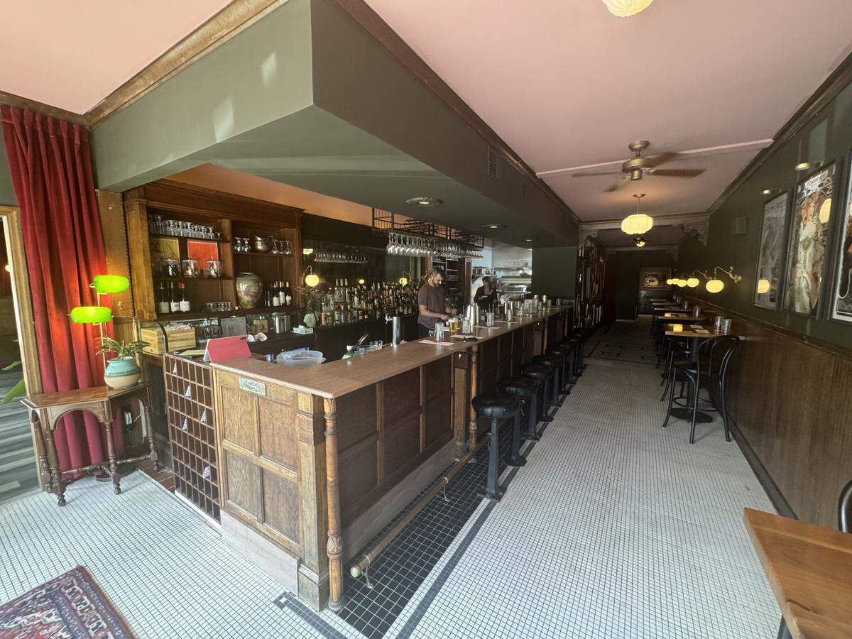 An interior view of a long, narrow upscale bar with a vintage aesthetic. It features dark wood paneling, a pink ceiling, and a long wooden bar with black stools. The floor is covered in small white and black mosaic tiles, and green shaded lamps provide a warm glow.