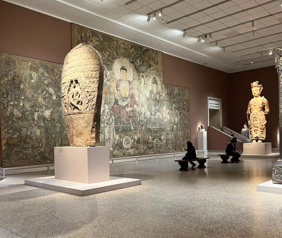 A large, spacious museum gallery with a high ceiling and a polished stone floor. The room features several large artifacts, including a tall, carved stone stele on a white pedestal in the foreground. In the background, a massive mural or fresco depicting a central Buddha figure surrounded by other deities covers a large wall. To the right, a tall, standing sculpture of a figure in robes is on display. Two visitors are seated on a bench in the middle of the gallery, observing the art.