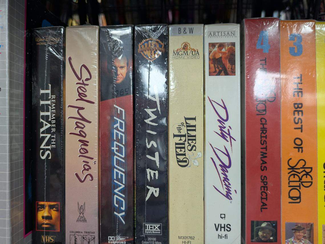 A close-up shot of a row of new, shrink-wrapped VHS tapes on a shelf, with their spines facing out. The titles visible from left to right are: Remember the Titans, Steel Magnolias, Frequency, Twister, Lilies of the Field, Dirty Dancing, a Christmas special, and The Best of Red Skelton.