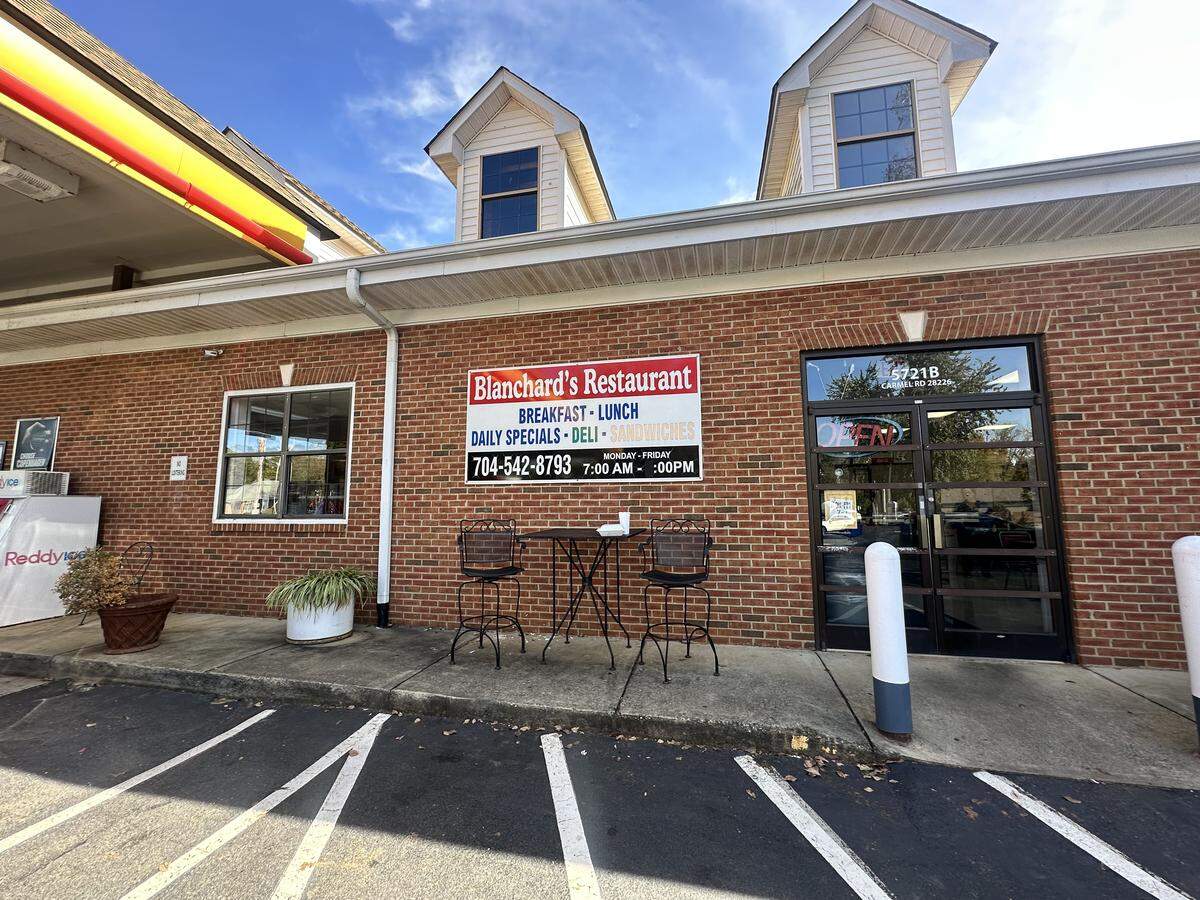 A straight-on, eye-level shot of the brick exterior of “Blanchard’s Restaurant” on a sunny day. The restaurant appears attached to a gas station, with a red and yellow canopy visible on the far left. A large white sign on the brick wall lists the restaurant’s name, its offerings (”BREAKFAST - LUNCH”, “DAILY SPECIALS - DELI - SANDWICHES”), phone number, and hours. A small black metal table and two chairs sit on the sidewalk in front, next to the glass entrance. The building is topped with a white upper level featuring two distinct dormer windows under a blue sky.
