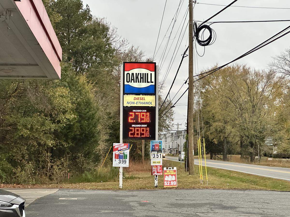 A wide, eye-level shot of a tall “OAKHILL” gas station price sign on an overcast day. The red, white, and blue sign displays red digital prices for “UNLEADED CASH” at “$2.79” and “UNLEADED CREDIT” at “$2.89”. The sign is located at the edge of a gas station, next to a utility pole and a two-lane road, with a background of trees in autumn colors.