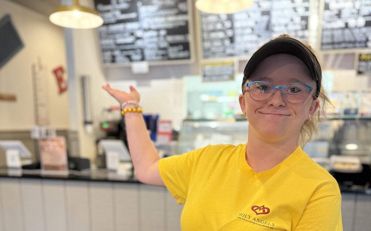 A person wearing a black visor, glasses, and a yellow t-shirt that says “Holy Angels” is smiling and gesturing to their right. They are standing behind a counter in a casual restaurant or cafe, with menu boards visible in the blurred background.