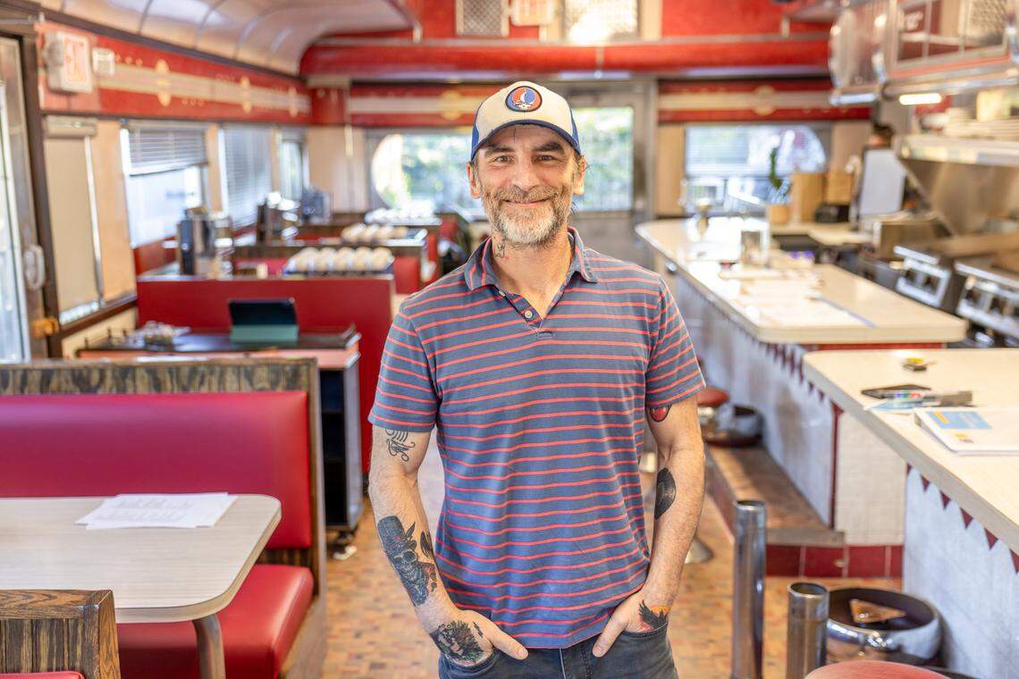 A medium shot of a smiling man with a grey beard and tattoos on his arms and hands, standing in the center of a vintage diner. He is wearing a blue and red striped polo shirt and a trucker hat with a circular logo. The diner’s interior features classic red booths, a long white counter with stools, and a curved metallic ceiling characteristic of a traditional railcar-style diner.