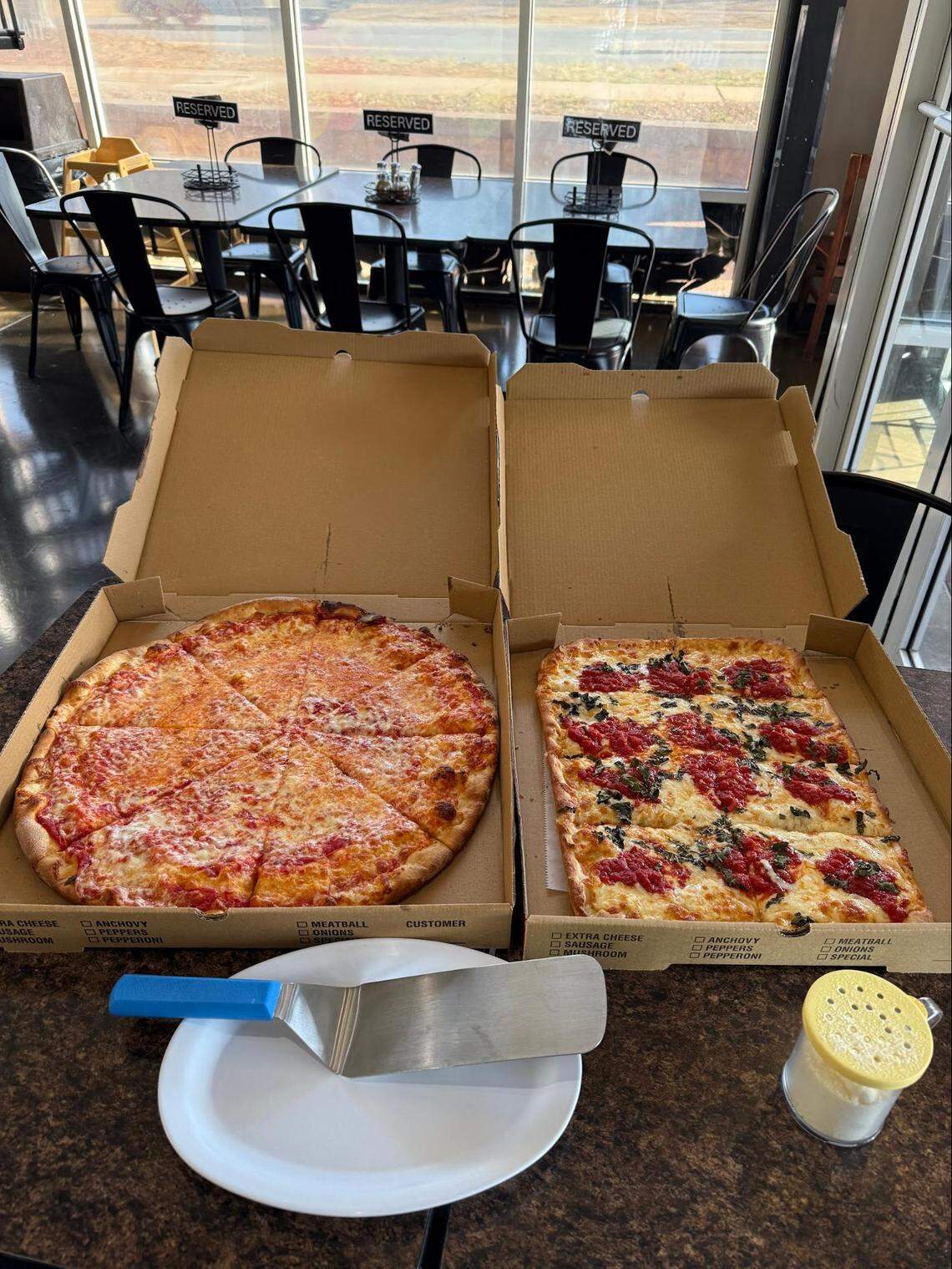 Two open pizza boxes sitting on a dark dining table. One contains a traditional round cheese pizza, while the other holds a rectangular “Grandma style” pizza topped with fresh tomato sauce and basil. A metal pizza server and a parmesan shaker are in the foreground.
