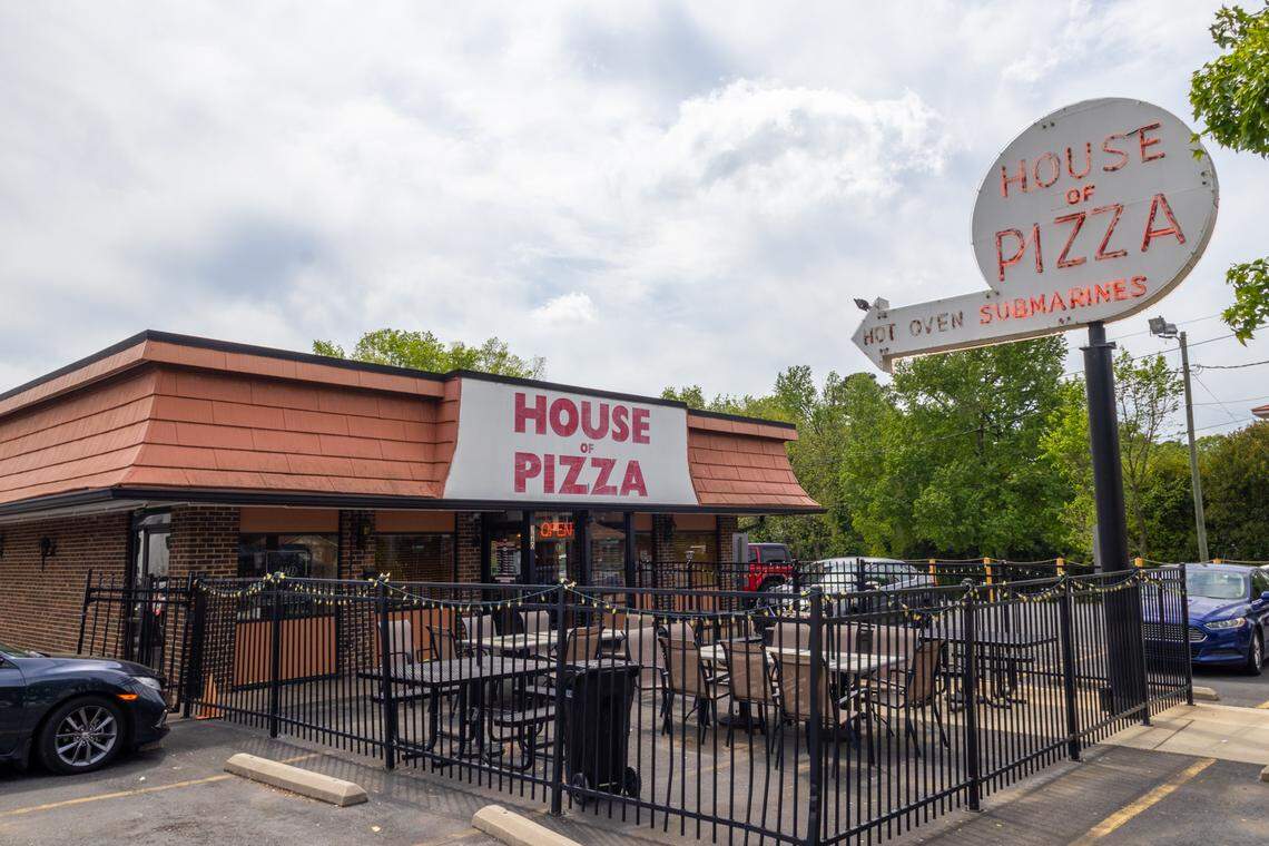 A wide, eye-level shot of the exterior of a ‘House of Pizza’ restaurant. The one-story brick building has a reddish-brown roof and a fenced-in outdoor patio with empty tables and chairs. A large sign on the building and a prominent, white, circular, retro-style sign on a pole both display the restaurant’s name in red letters. The pole sign also has an arrow pointing to the entrance, advertising ‘HOT OVEN SUBMARINES.’ The scene is set under a partly cloudy sky with green trees in the background.