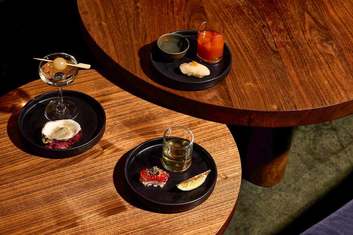 An overhead view of two circular wooden tables displaying three “perfect pairings.” Each pairing is served on a small matte black plate and includes a single bite of food—an oyster on the half shell, a piece of tuna nigiri, and a piece of yellowtail nigiri—each accompanied by a specific cocktail or spirit in various glassware.