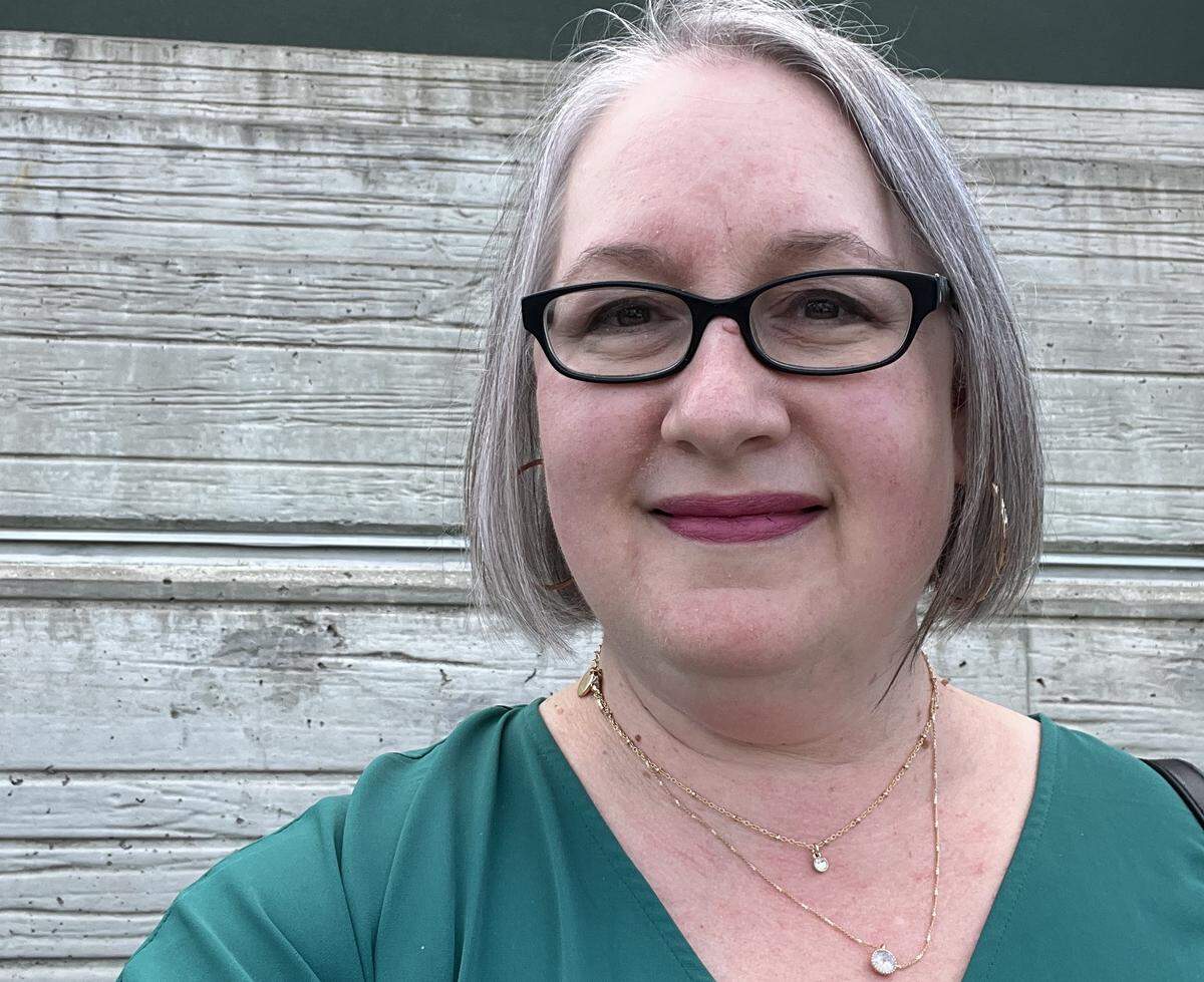 A close-up selfie of a smiling person with short, silver-gray hair and black-framed glasses wearing a bright emerald green top and two delicate gold chain necklaces, one with a small clear crystal and one with a larger pendant. The person’s makeup includes pink lipstick. The background is a slightly weathered, light gray-blue wooden wall with horizontal planks.