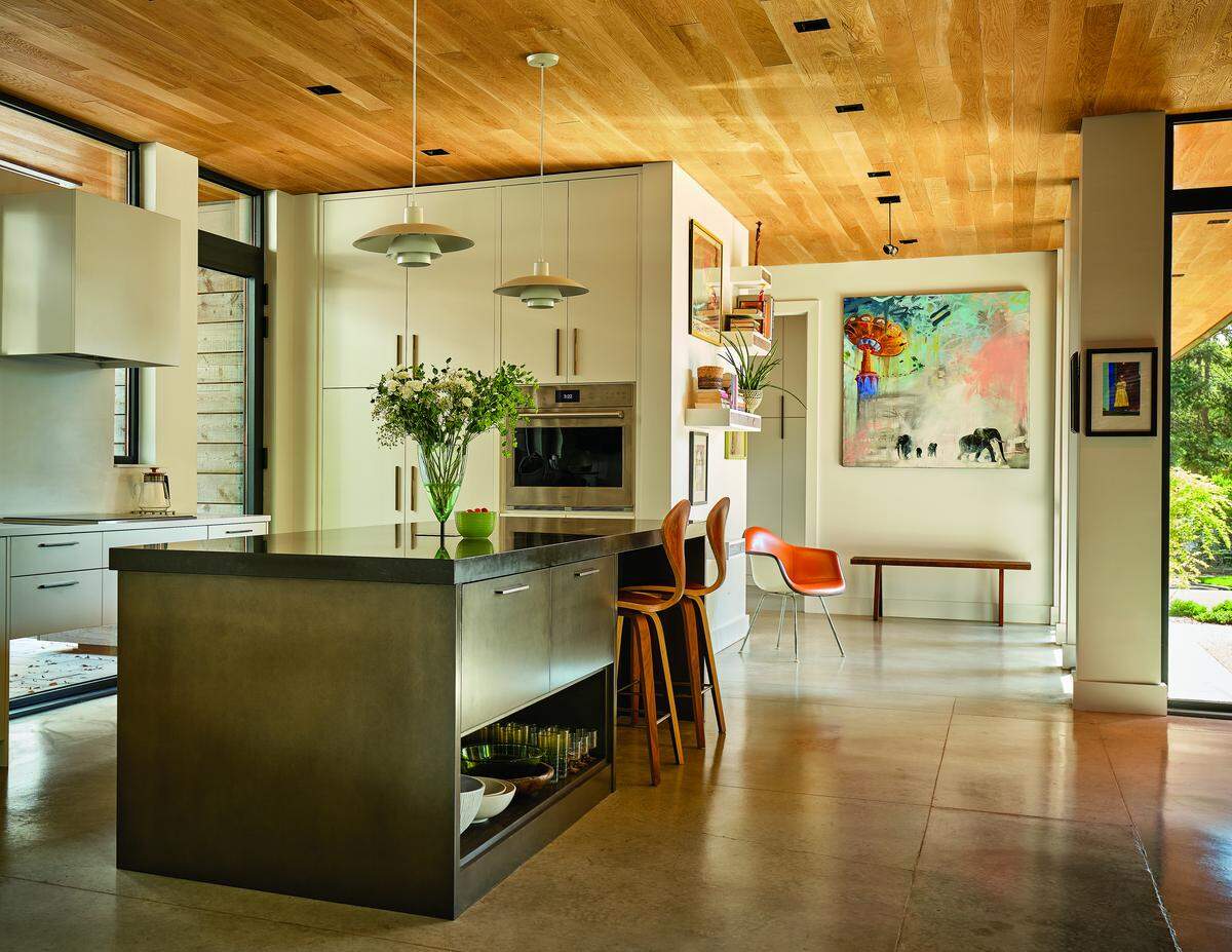 A wide shot of a bright, modern kitchen with mid-century modern influences. The room features a warm wooden ceiling and polished concrete floors. A large, dark-topped kitchen island with open shelving sits in the center, complemented by two orange bar stools. Behind the island, a wall of minimalist white, flat-panel cabinets contains a built-in oven and is lit by two white pendant lights. To the right, an entryway is decorated with a large, colorful abstract painting of elephants. Large windows and glass doors let in abundant natural light.