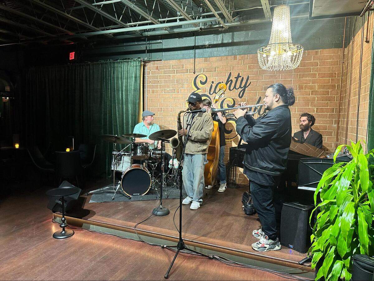 A five-piece jazz band is performing on a low wooden stage in a dimly lit club. In the foreground, a trumpeter plays, while other musicians are on saxophone, drums, upright bass, and piano. They are performing in front of a brick wall with a logo that says “Eighty Eights” in cursive, and a crystal chandelier hangs from the ceiling. A large green plant frames the right side of the image.