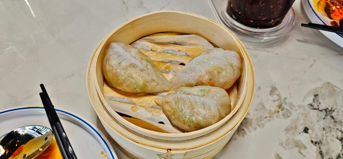 A close-up of three translucent, crescent-shaped steamed dumplings served in a round bamboo steamer basket on a restaurant table.