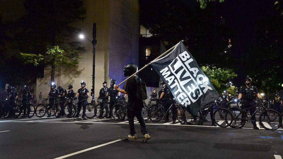 A protester carries a Black Lives Matter flag as he skateboards in front of Charlotte Mecklenburg police officers along College Street in Charlotte, NC on Saturday, August 22, 2020.