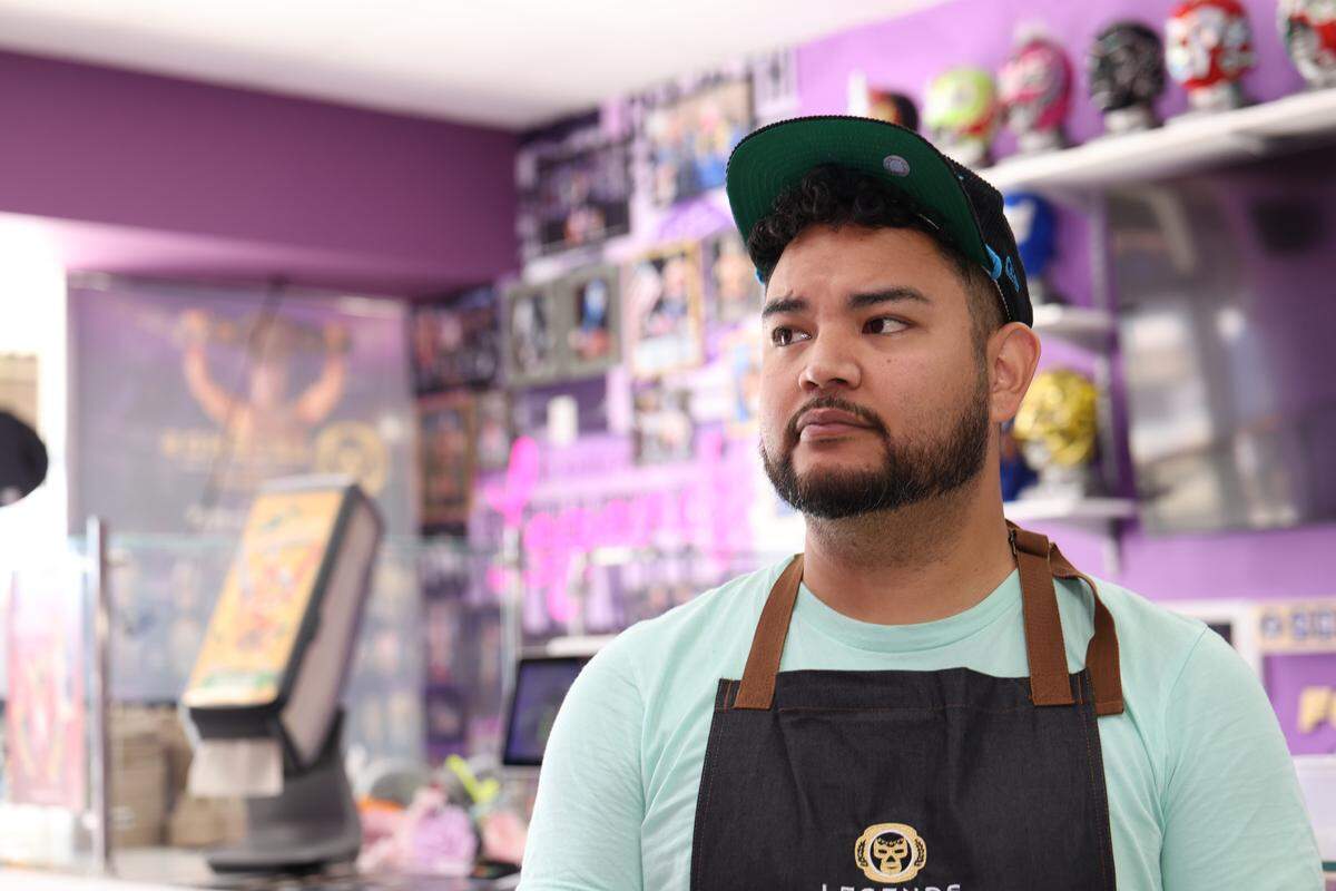 A tight, close-up shot of an ice cream shop owner wearing a mint-colored t-shirt, apron, and a backward green baseball cap, looking off to the right. The background is blurred, revealing a purple wall with framed photos and a bright pink neon sign.
