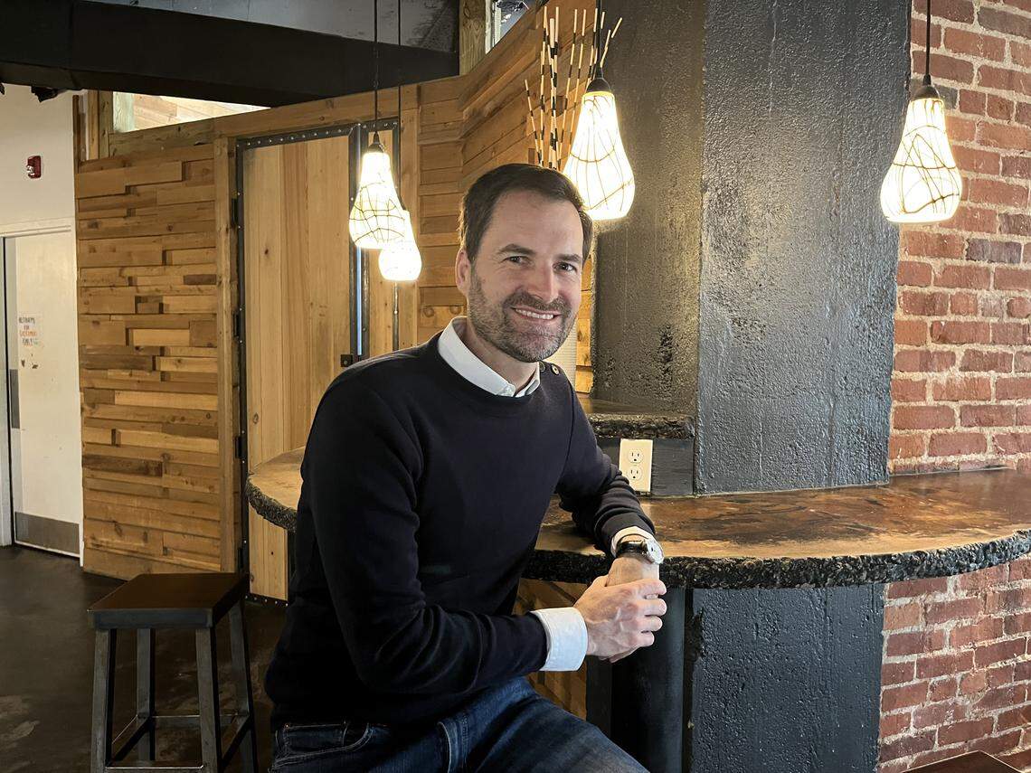 A medium, eye-level shot of a smiling person, wearing a blue sweater and collared shirt, sitting at a curved bar counter. He is in a warmly-lit restaurant, positioned between an exposed red brick wall on the right and a modern, wood-paneled wall on the left, with several stylish pendant lights hanging overhead.