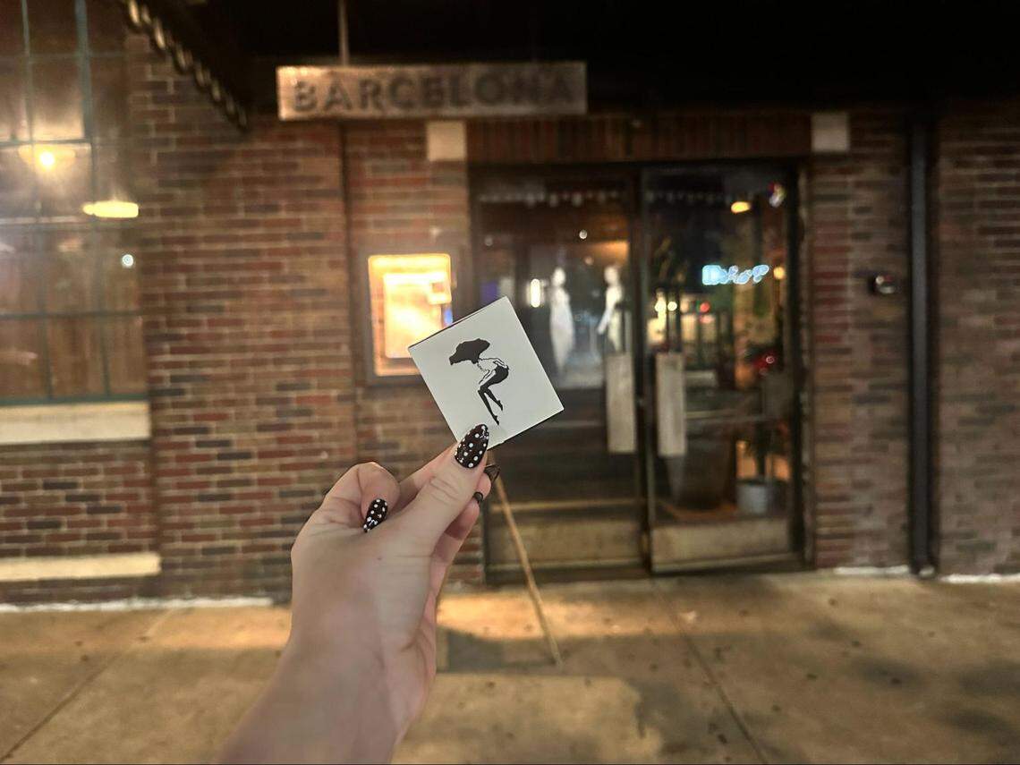 A person’s hand, adorned with black and white polka-dot nail art, holds up a white matchbook in front of a restaurant at night. The matchbook features a black silhouette of a woman holding an umbrella. The background shows the warmly lit, brick exterior of the restaurant, with a sign that reads “BARCELONA” above the entrance.